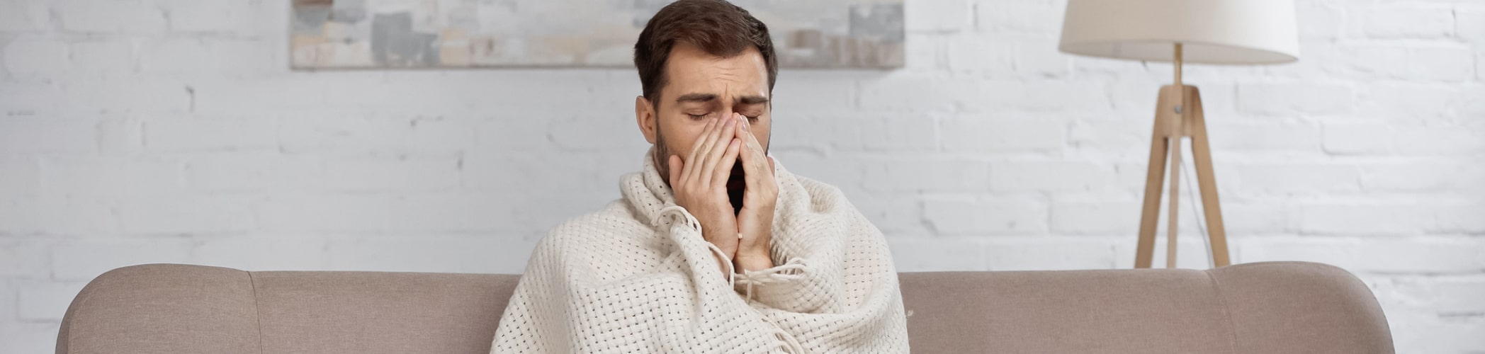 Van welke lichamelijke klachten wil jij graag af? Afbeelding van een man die op de bank zit met een deken om zich heen die duidelijk ziek is en met zijn handen voor zijn mond zit. Achter hem staat een lamp.
