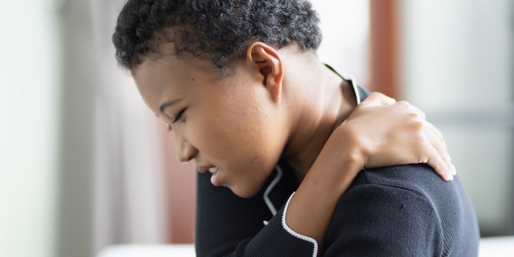Fibromyalgie testen Een foto van een donkere vrouw met kort zwart haar die haar schouder omklemt omdat ze pijn heeft. De fot o