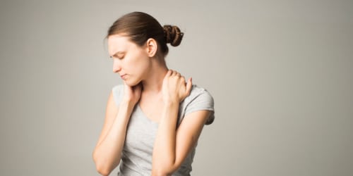 Hoe kan je fibromyalgie ontdekken_ Een afbeelding van een vrouw met een beige tshirt aan die haar handen op de pijnlijke spieren van haar nek heeft liggen. Ze wil weten hoe ze fibromyalgie kan ontdekken