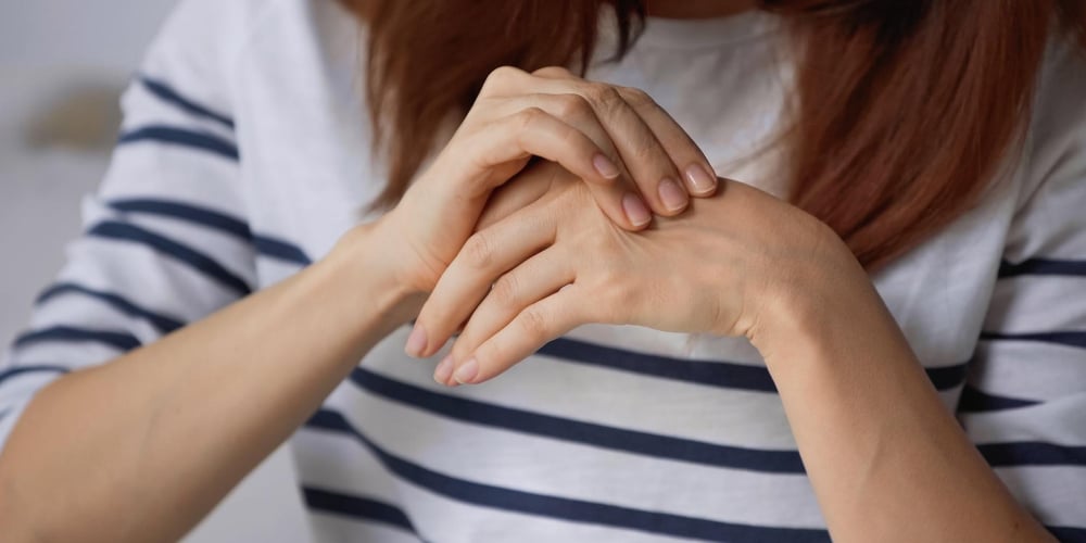 Hoe kan je reuma ontdekken_ Vrouw in een wit blauwe trui die haar zere hand vasthoudt. De foto symboliseert hoe je reuma kan ontdekken
